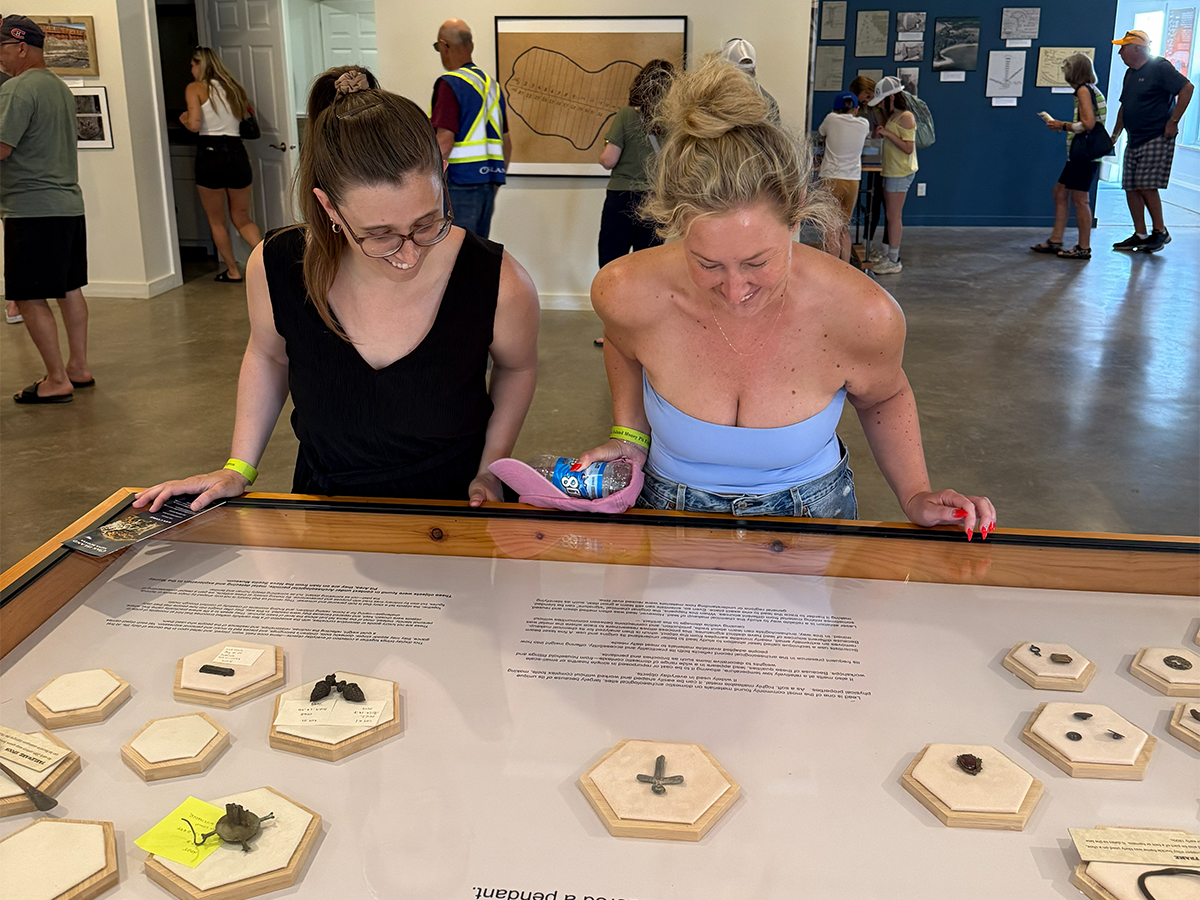 Visitors examining a display case of small Oak Island artifacts, with the large island map on the wall behind them