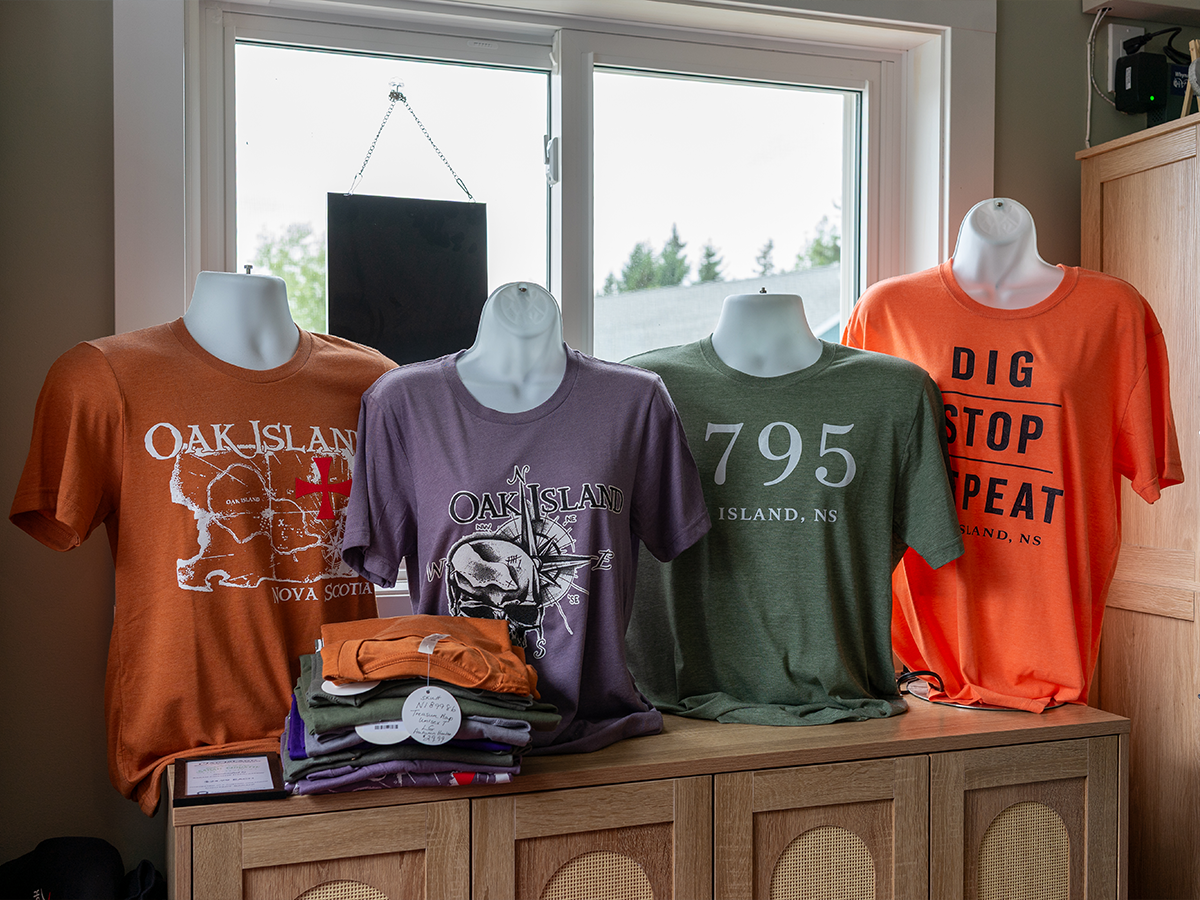 A display of Oak Island t-shirt designs — Oak Island NS map, skull-and-compass, “1795”, and “Dig · Stop · Repeat”